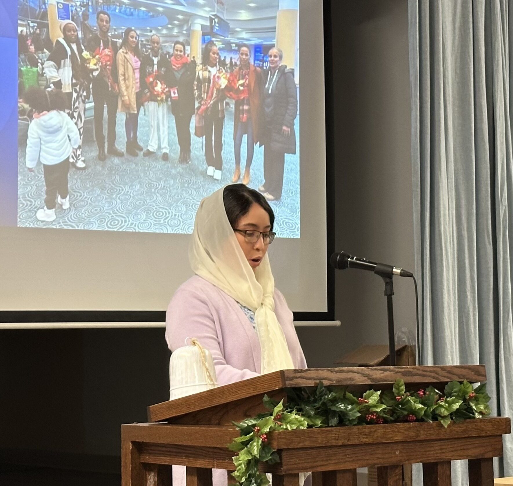 A woman in a pink shirt wearing a white headscarf stands behind a brown podium speaking into a microphone.