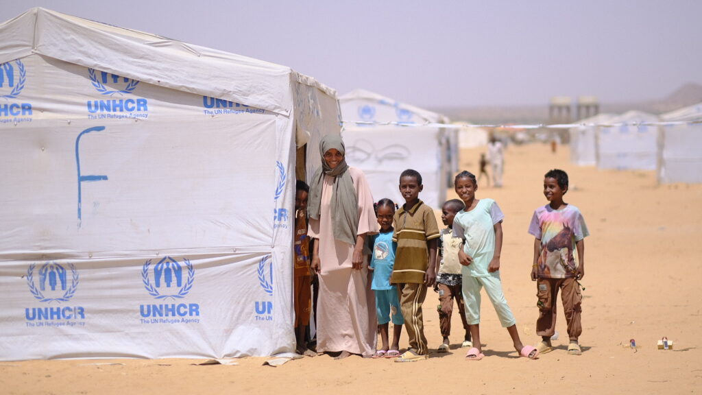 A family stands outside of a temporary shelter with the UNCHR logo on it
